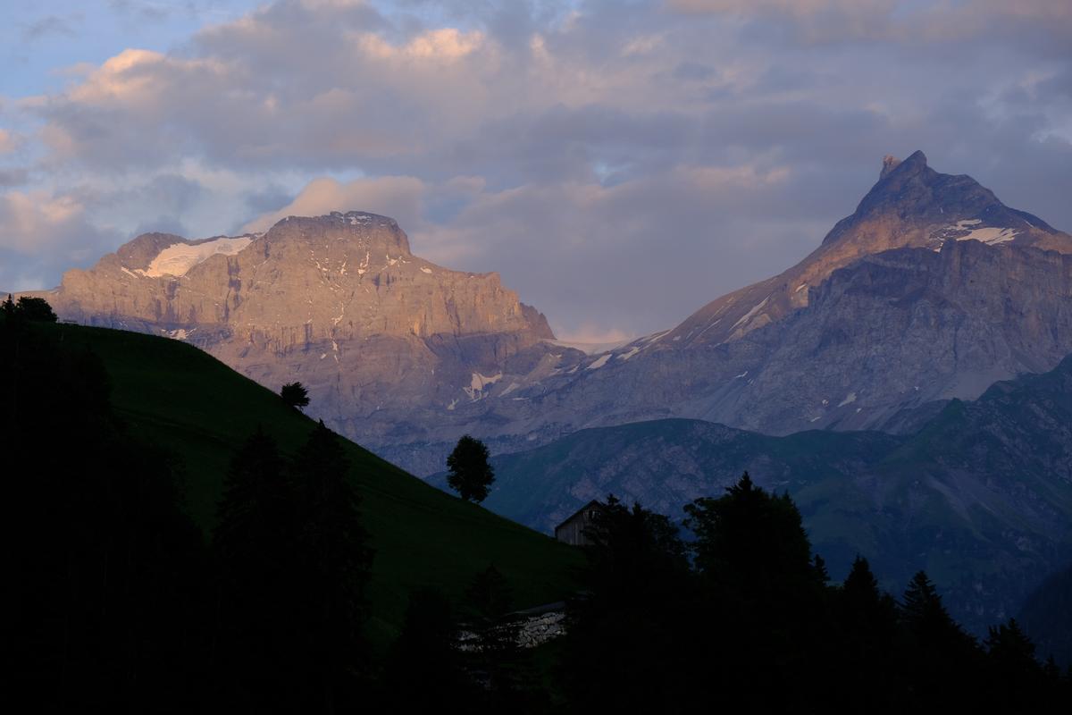 A hill in the foreground is silhouetted against mountains lit by the evening sun, Switzerland.