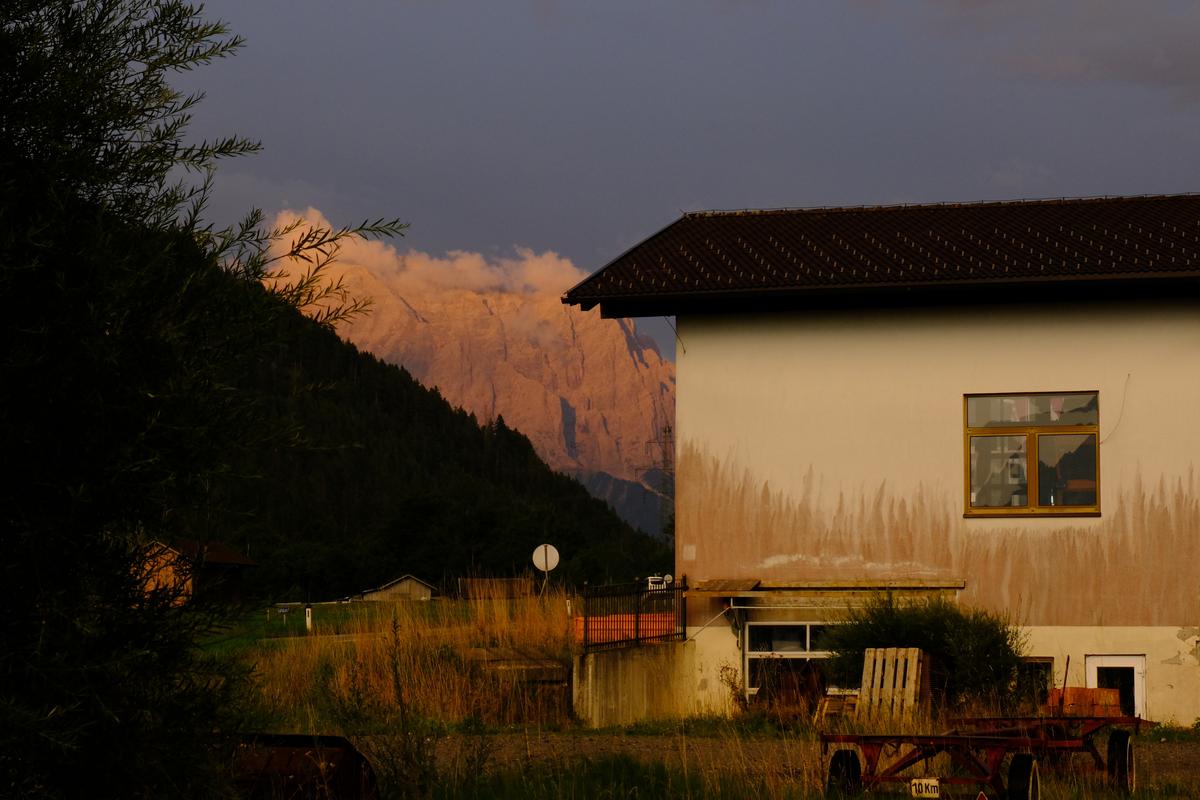 A white house in the foreground against a darkening sky as the setting sun lights the mountain behind. Austria