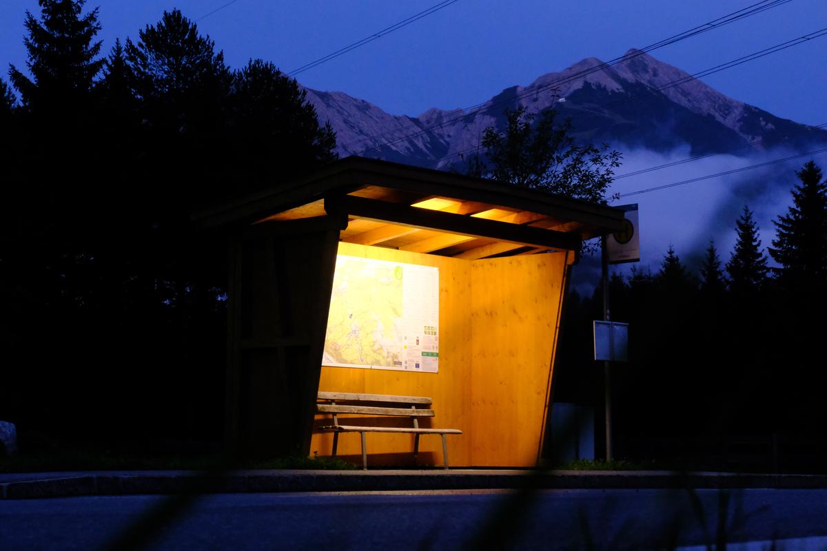 A brightly lit rural bus stop at night in Austria, mountains form a backdrop