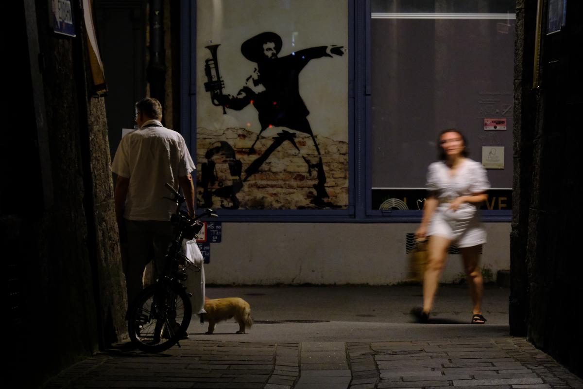 A woman entering the alley from the right is a blur while a man, a dog, and a bicycle are still to the left. Behind them, a mock depiction of a revolutionary throwing a trumpet in a Banksy-esque style