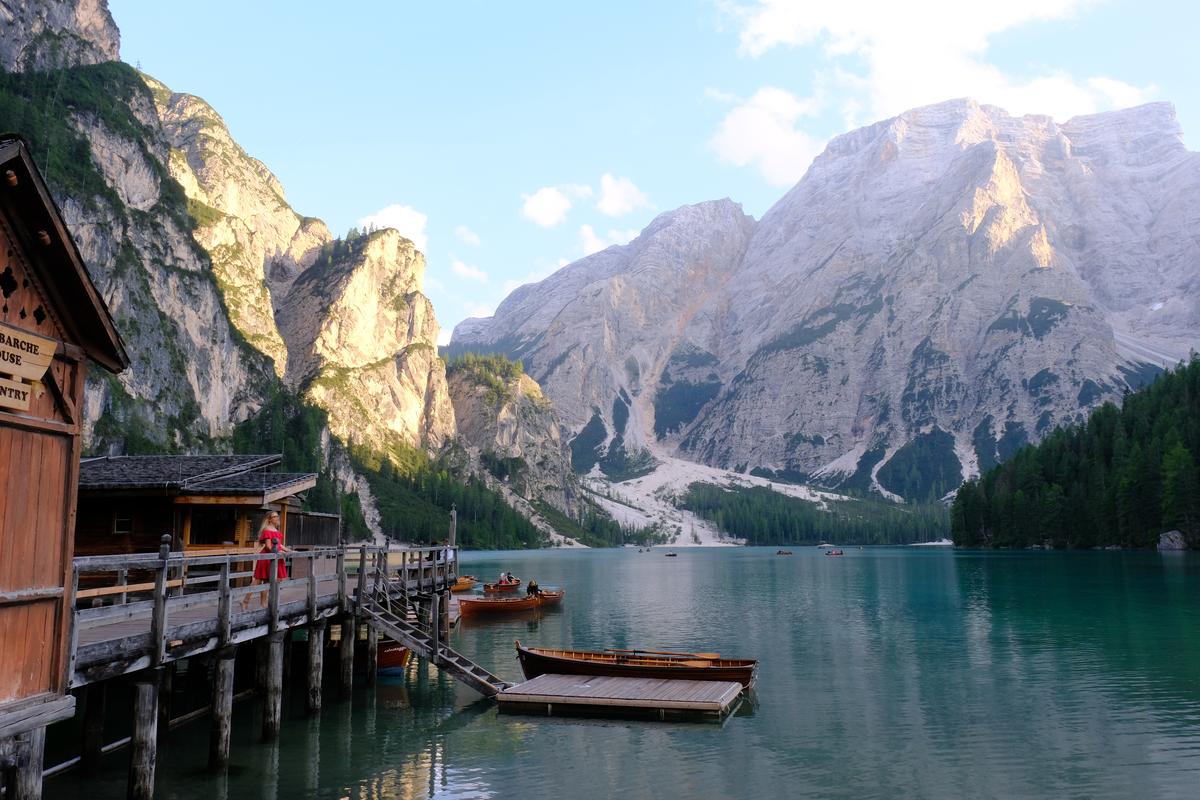 A view over Lago di Braies, Italy. Rowing boats are visible on the far side of the lake, to the left a woman in a red dress leans against the railing of the dock.