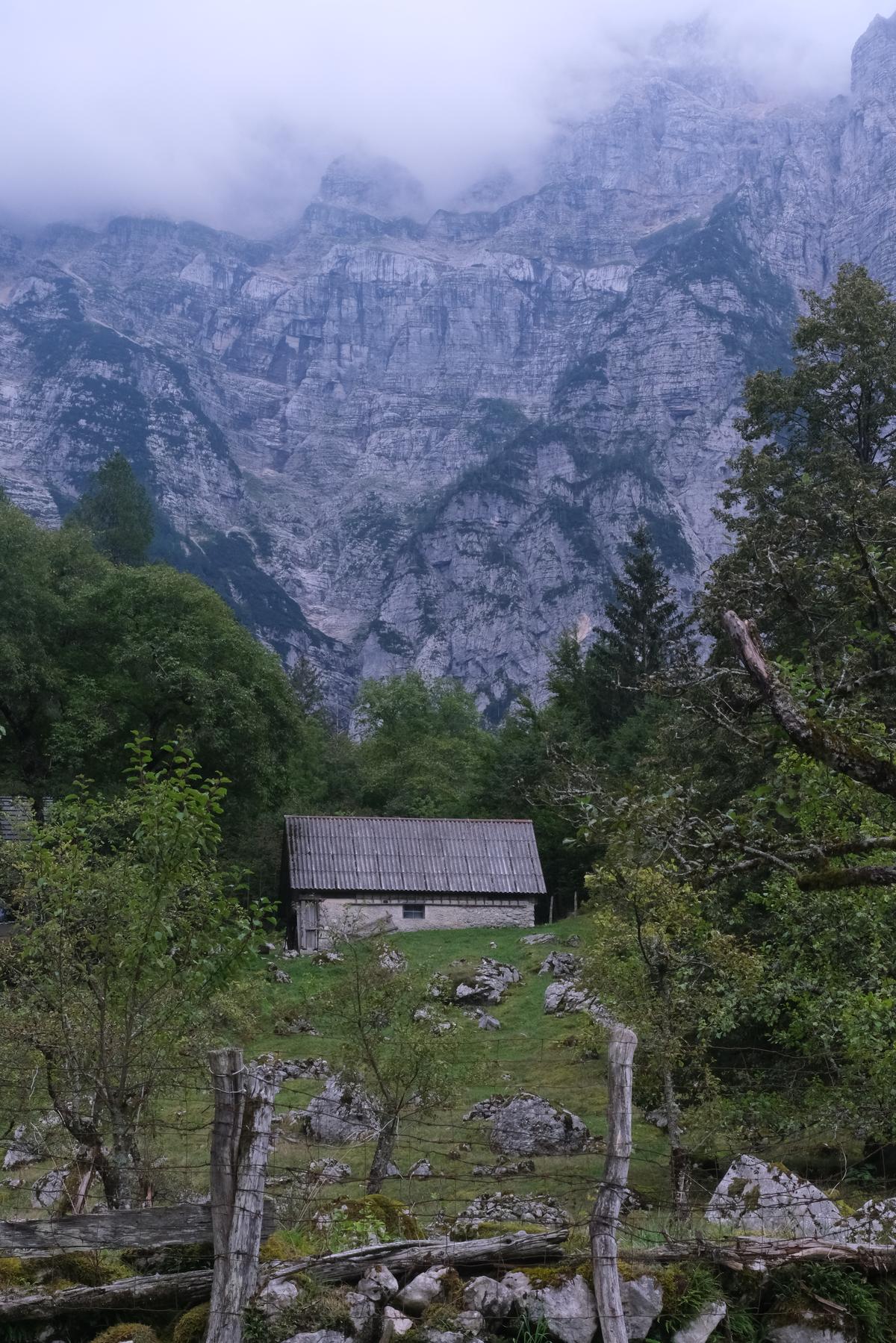 A stone outhouse nestled in the trees behind a barbed wire fence. Behind, the Slovenian Alps reach up into the clouds. 