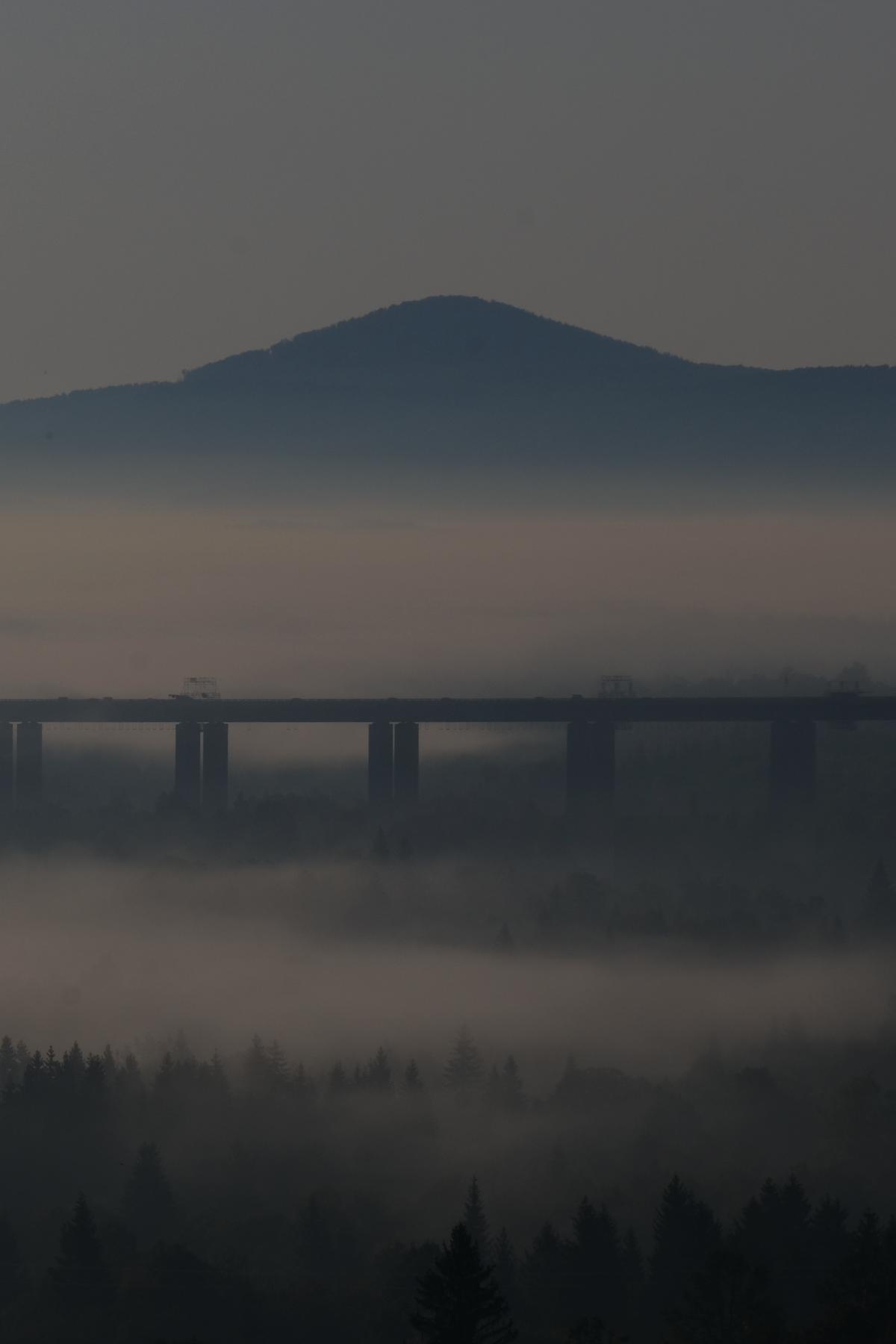 A forested valley in low cloud, backdropped by a tree covered mountain, bisected by a massive highway bridge. Croatia