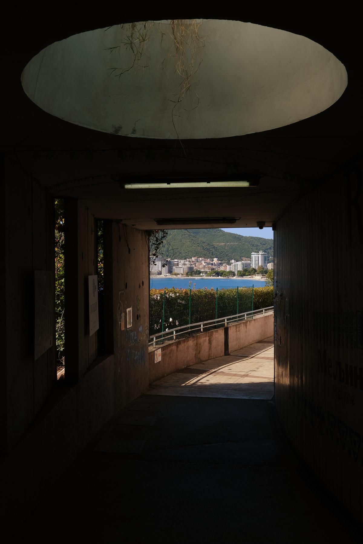 A pedestrian tunnel along the coast, dimly lit by a skylight with hanging vegetation, between Sveti Stefan and Budva, Montenegro.