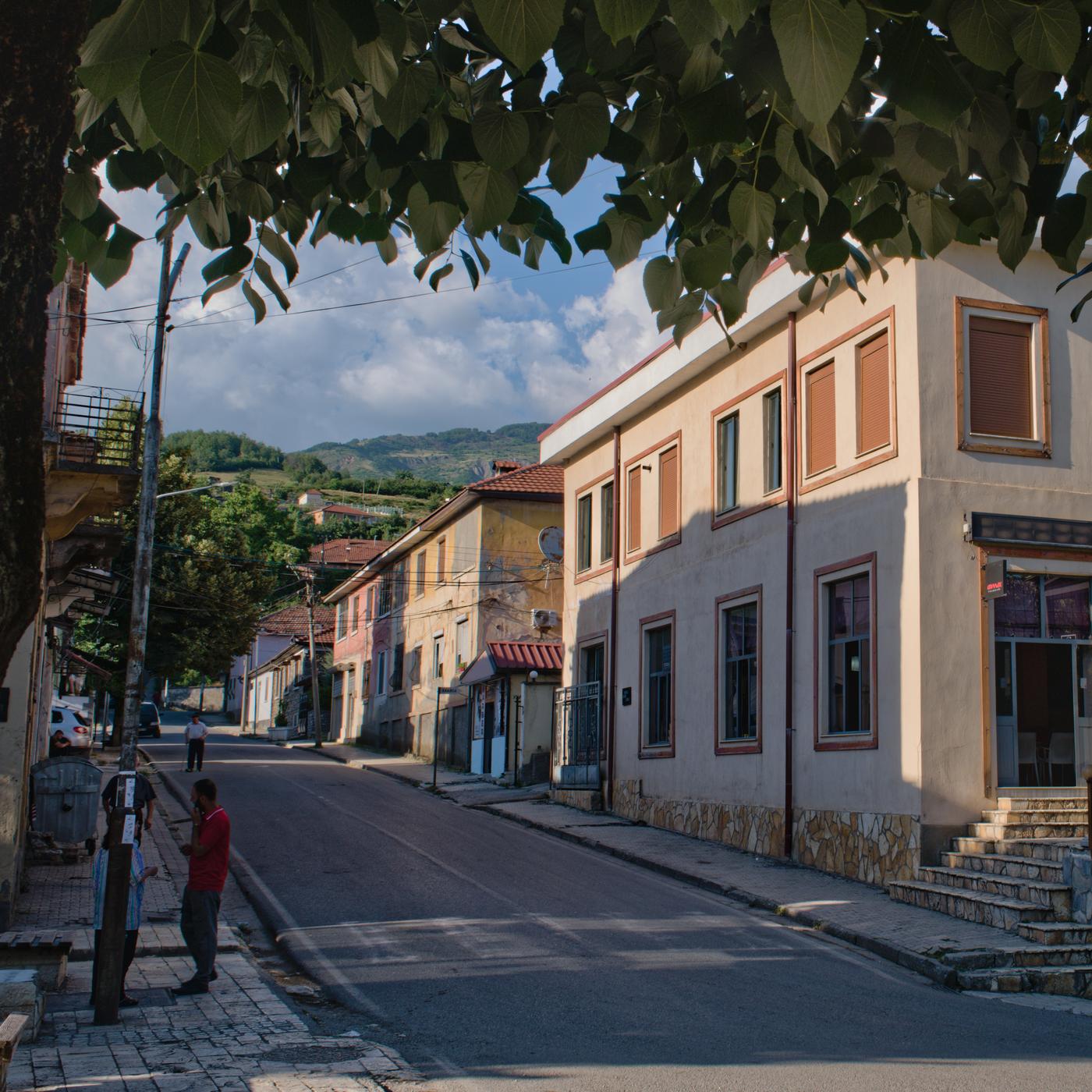 The falling evening light catches the upper floors of several buildings while a handful of pedestrians occupy the shade on a street in Peshkopi, Albania