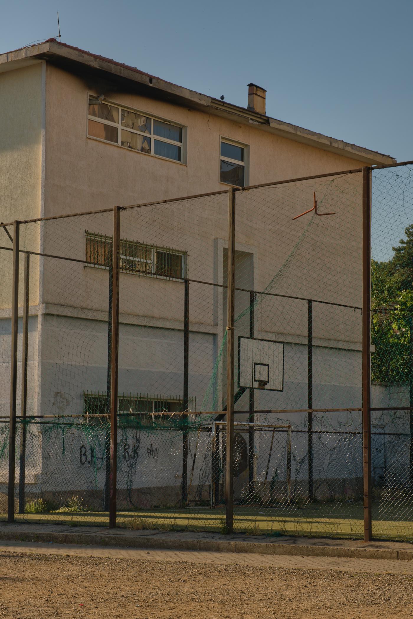 A graffitied wing of a school behind a chain link fenced basket ball court. Peshkopi, Albania