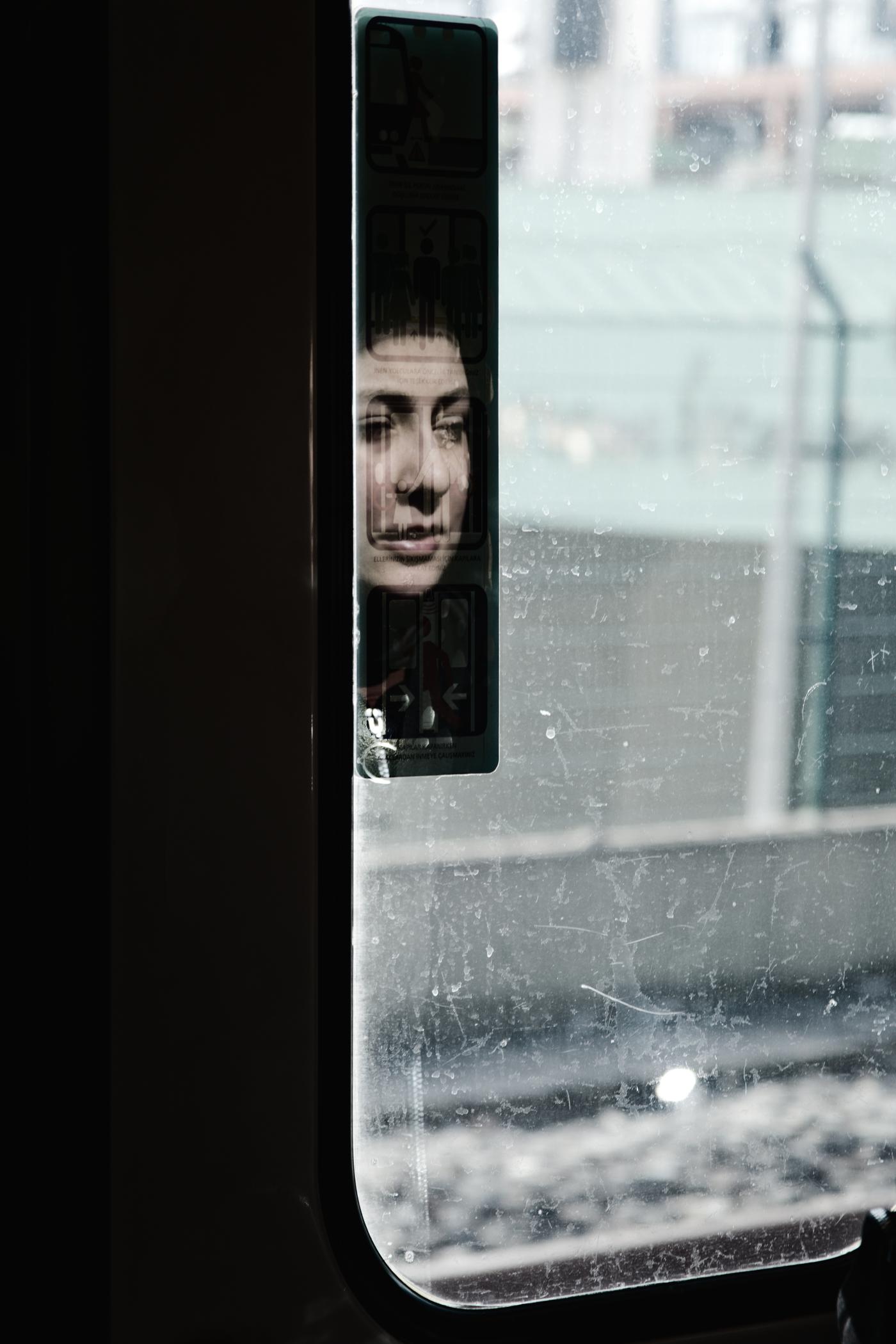 A young woman’s face catches in the reflection of a train window, made vivid by an emergency exit sign. Istanbul, Turkey