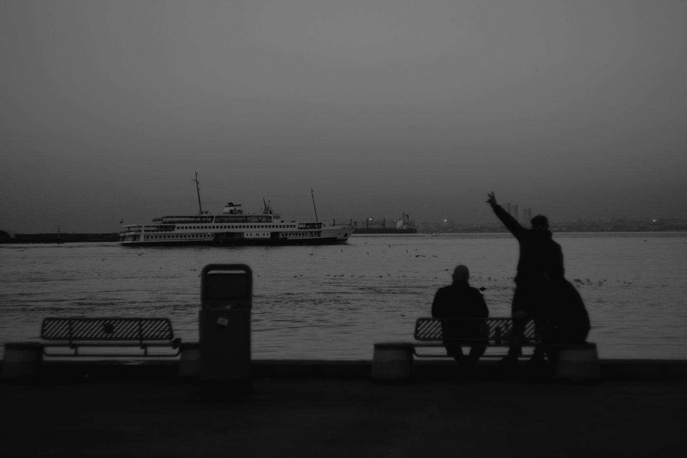 A man, silhouetted against the sea and sky, makes the peace sign. Beyond, a ferry is crossing the Bosphorus. Istanbul, Turkey