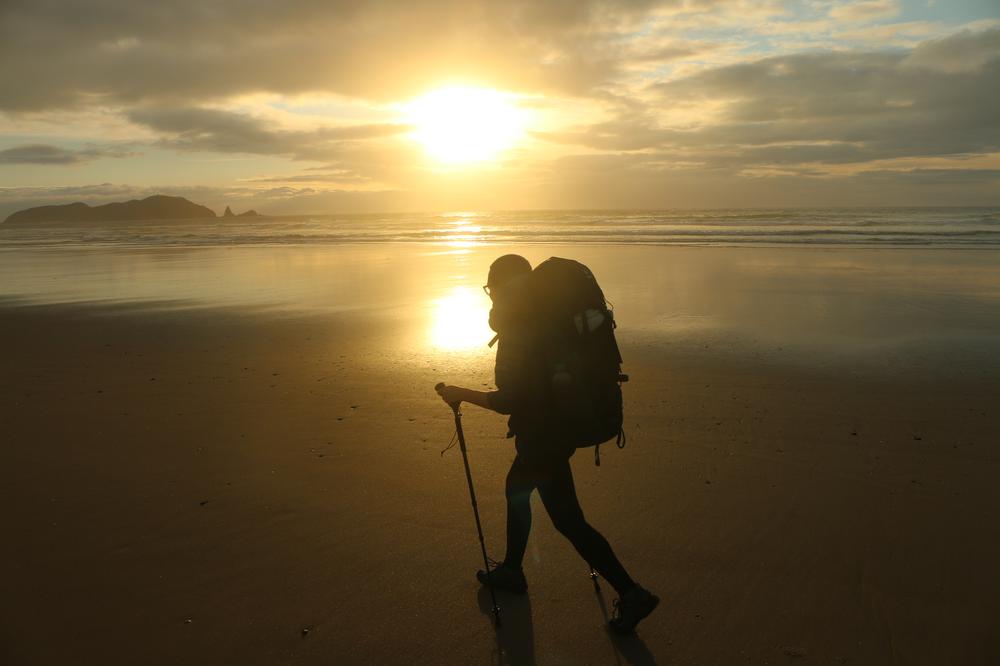 Rose crosses Te Werahi beach as the sun sets on the water behind her.