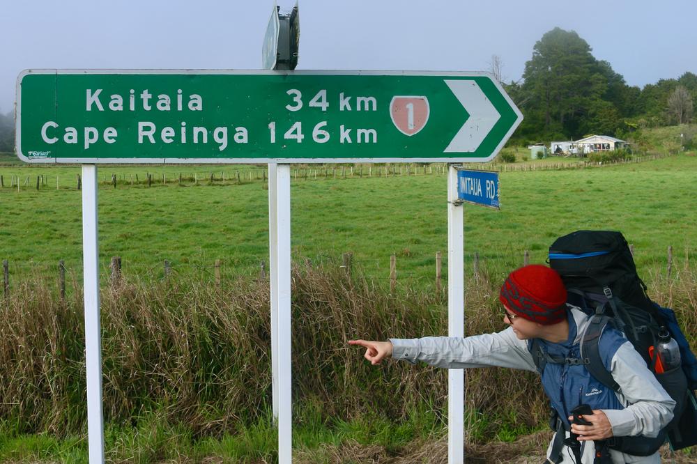 Rose points south in front of roadsign showing the distance back to Cape Reinga to the north.