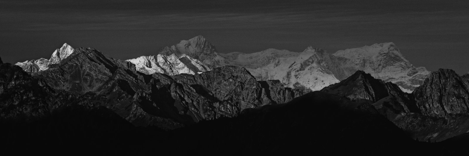 Clouds cast dark shadows over the mountains while a narrow band of sunlight hits a ridge in the middle distance. Western Austria.