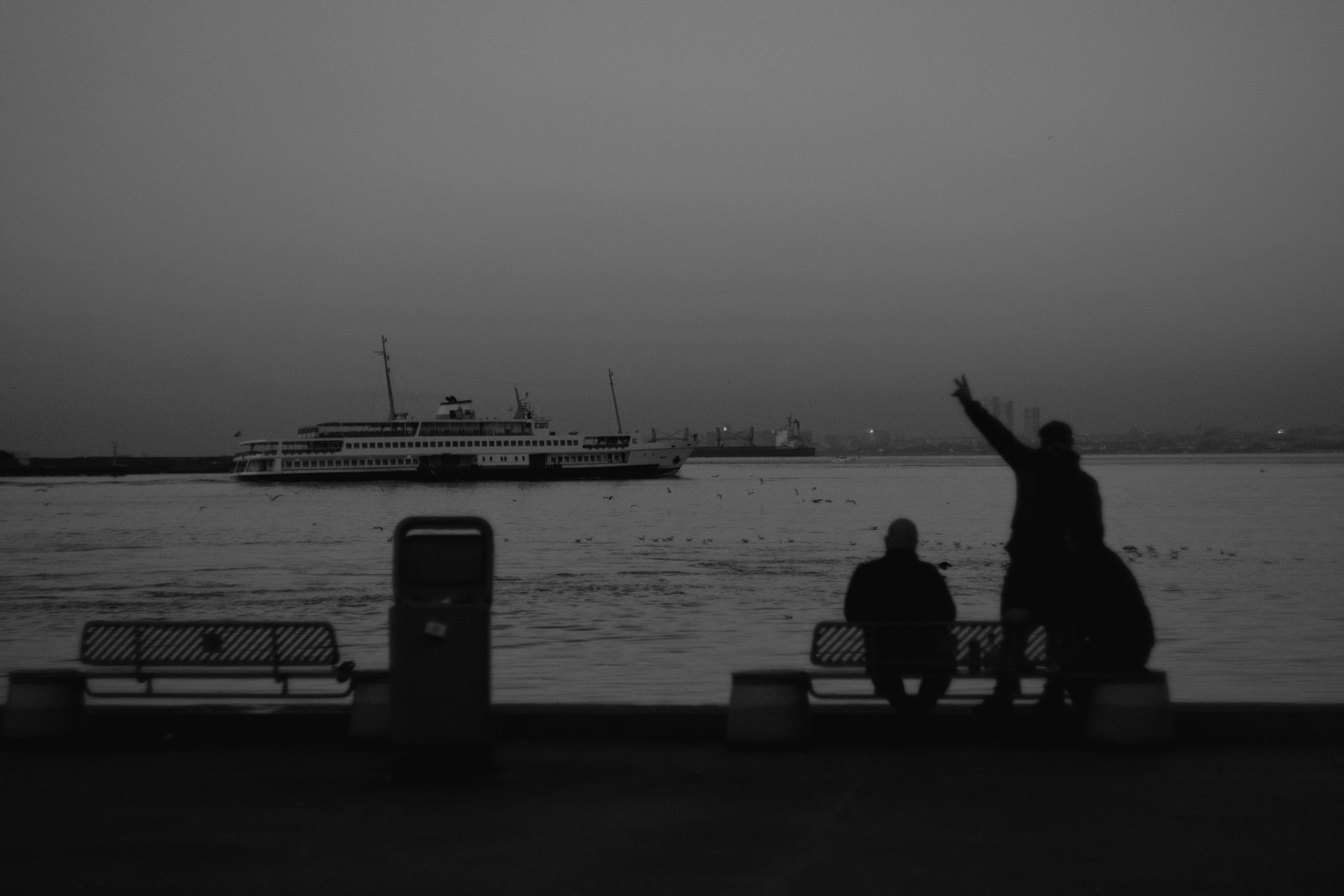 A man, silhouetted against the sea and sky, makes the peace sign. Beyond, a ferry is crossing the Bosphorus. Istanbul, Turkey