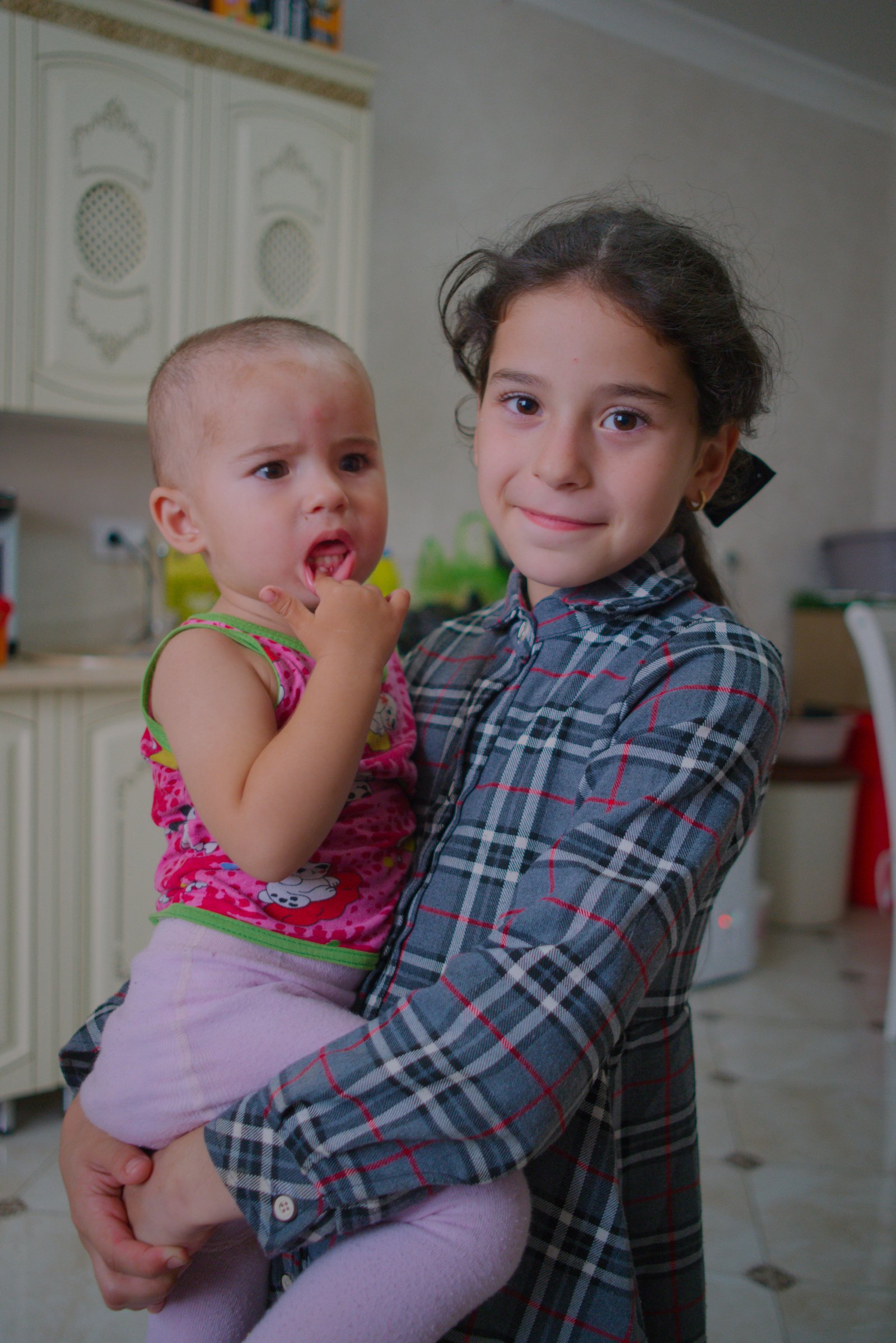 Hawa (Хава) holds her younger sister and watches me eat the food her parents have laid out for me. Chechnya, Russia