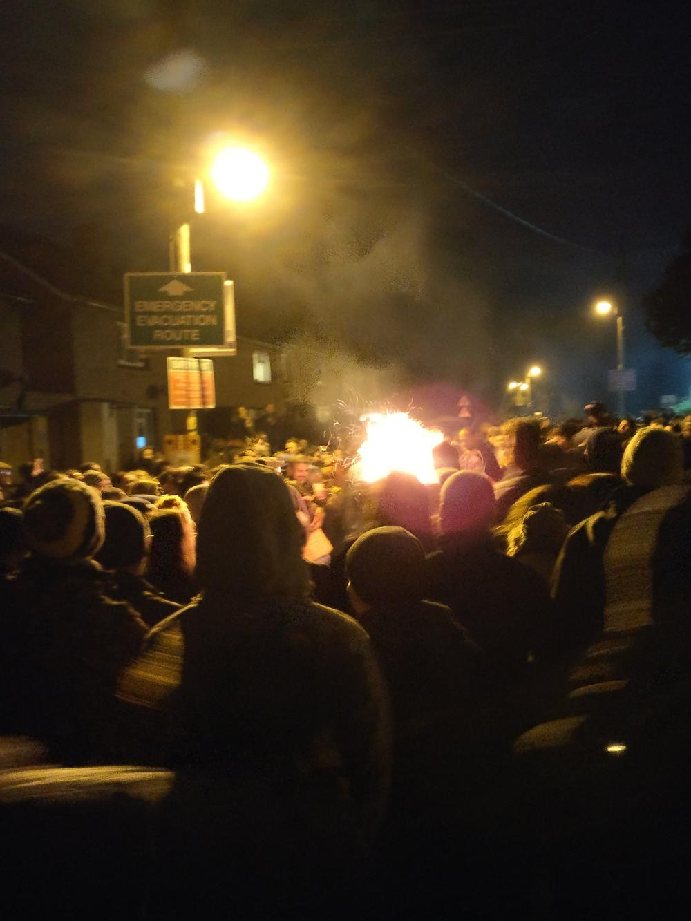 A crowd gathers around a burning barrel in Ottery St Mary